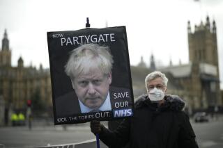 FILE - An anti-Conservative Party protester holds a placard with an image of British Prime Minister Boris Johnson including the words "Now Partygate" backdropped by the Houses of Parliament, in London, Wednesday, Dec. 8, 2021. British police are getting ready to issue a first batch of fines on over parties held by Prime Minister Boris Johnson’s staff during coronavirus lockdowns. The Metropolitan Police declined to confirm reports multiple U.K. media outlets that fines would come as soon as Tuesday, March 29, 2022, saying it would not give “a running commentary” on its probe. (AP Photo/Matt Dunham, File)