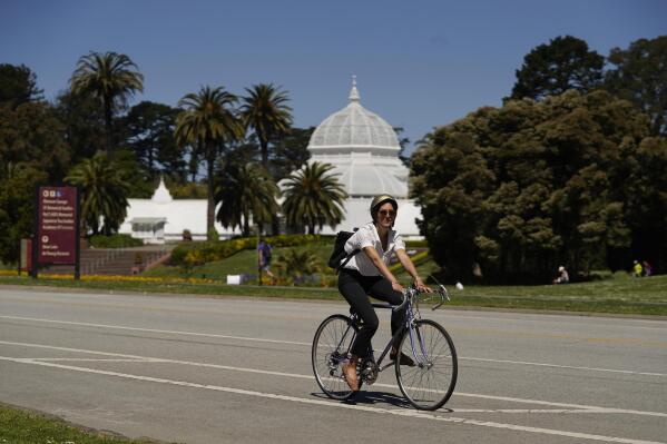 A woman on a bicycle rides along the car-free John F. Kennedy Drive in Golden Gate Park with the Conservatory of Flowers in the background, Wednesday, April 28, 2021, in San Francisco. At the start of the pandemic, San Francisco closed off parts of a major beachfront highway and Golden Gate Park to cars so that people had a safe place to run and ride bikes. Open space advocates want to keep those areas car-free as part of a bold reimagining of how U.S. cities look. But opponents decry the continued closures as elitist, unsafe and nonsensical now that the pandemic is over and people need to drive again. (AP Photo/Eric Risberg)