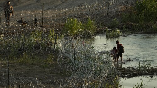 A Texas state trooper watches as young migrants walk along concertina wire on the banks of the Rio Grande as they try to enter the U.S. from Mexico in Eagle Pass, Texas, Thursday, July 6, 2023. Texas Republican Gov. Greg Abbott has escalated measures to keep migrants from entering the U.S. He's pushing legal boundaries along the border with Mexico to install razor wire, deploy massive buoys on the Rio Grande and bulldozing border islands in the river. (AP Photo/Eric Gay)