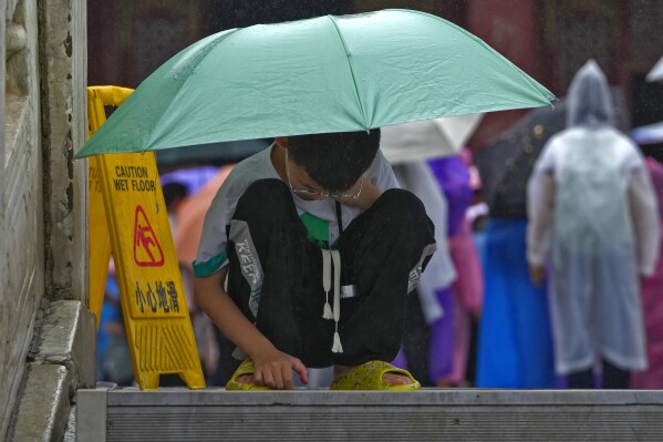 A boy covering himself with an umbrella from the rain, browses a smartphone placed on the ground at the Forbidden City in Beijing on July 13, 2023. China's internet watchdog has laid out regulations to curb the amount of time children spend on their smartphones, in the latest blow to firms such as Tencent and ByteDance, which run social media platforms and online games. (AP Photo/Andy Wong)