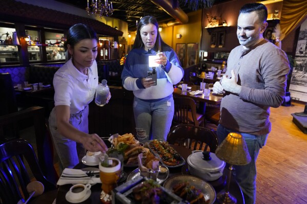 Restaurant owner Ana Acela Perez, from left, assists content creator Jensen Savannah, and her fiance and brand manager Jorge Millares, with a video for her business at El Puro Cuban Restaurant, Thursday, March 14, 2024, in Charlotte, N.C. (AP Photo/Erik Verduzco)