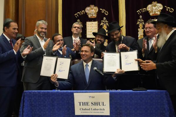 Surrounded by state legislators and Jewish leaders, Florida Gov. Ron DeSantis, center, holds up two bills that he signed, Monday, June 14, 2021, at the Shul of Bal Harbour, a Jewish community center in Surfside, Fla. DeSantis visited the South Florida temple to denounce anti-Semitism and stand with Israel, while signing a bill into law that would require public schools in his state to set aside moments of silence for children to meditate or pray. (AP Photo/Wilfredo Lee)