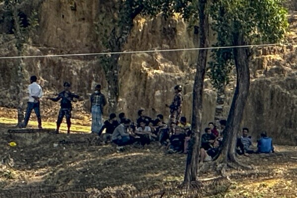 Members of Myanmar Border Guard Police, in civilian clothing, sit under the shade of trees after abandoning their posts following an alleged attack by members of the Arakan Army as Bangladesh border guards stand guard in Ghumdhum, Bandarban, Bangladesh, on Monday, Feb. 5, 2024. Nearly a hundred members of Myanmar's Border Guard Police have fled their posts and taken shelter in Bangladesh during fighting between Myanmar security forces and an ethnic minority army, an official of Bangladesh's border agency said Monday. (AP Photo/Shafiqur Rahman)