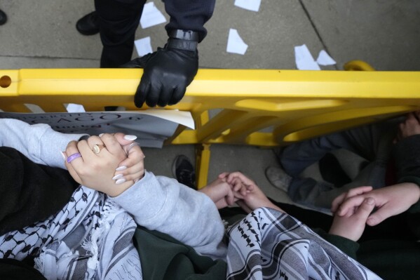 Palestīniešu atbalstošie demonstranti saista rokas un saspiež plaukstas, kamēr Čikāgas Universitātes policists turas pie barikādes, kad policisti bloķē protestētājus no universitātes laukuma studentu nometnes demontāžas laikā, otrdien, 2024. gada 7. maijā, Čikāgā.  (AP foto/Čārlzs Rekss Arbogasts)