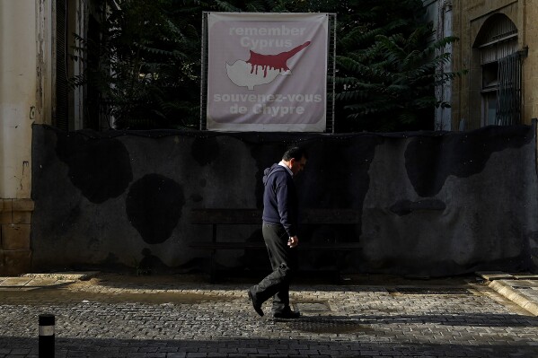 A man walks across the U.N buffer zone in front of a blocked road as a banner shows the Cyprus island divided, the Turkish occupied area at the north and Cyprus republic at the south, in divided capital Nicosia, Cyprus, on Wednesday, May 15, 2024. Chances of restarting formal talks to mend Cyprus' decades-long ethnic division appeared dimmer as the leader of the breakaway Turkish Cypriots told a United Nations envoy that he saw no common ground with Greek Cypriots for a return to negotiations. (AP Photo/Petros Karadjias)
