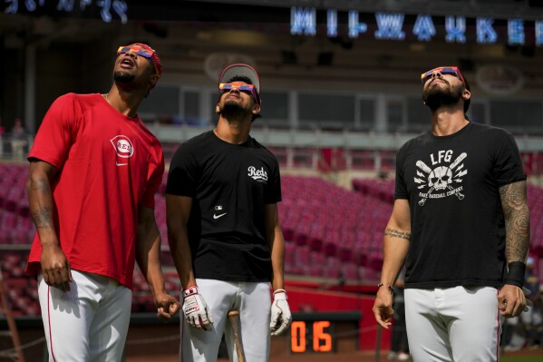 Cincinnati Reds' Will Benson, left, Bubba Thompson, middle, and Jonathan India, right, use special glasses as they watch the sun during a solar eclipse before a baseball game against the Milwaukee Brewers in Cincinnati, Monday, April 8, 2024. (AP Photo/Aaron Doster)