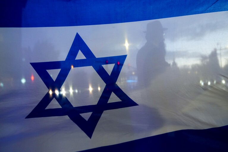 A demonstrator crossing the street is silhouetted behind a flag of Israel during a rally in support of Israel Monday, Oct. 9, 2023, in Bellevue, Wash. (AP Photo/Lindsey Wasson, File)