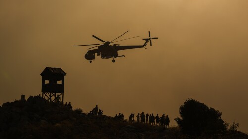 A firefighting helicopter flies through smoke as people watch, at the fire in Mandra west of Athens, on Tuesday, July 18, 2023. In Greece, where a second heatwave is expected to hit Thursday, three large wildfires burned outside Athens for a second day. Thousands of people evacuated from coastal areas south of the capital returned to their homes Tuesday when a fire finally receded after they spent the night on beaches, hotels and public facilities. (AP Photo/Petros Giannakouris)