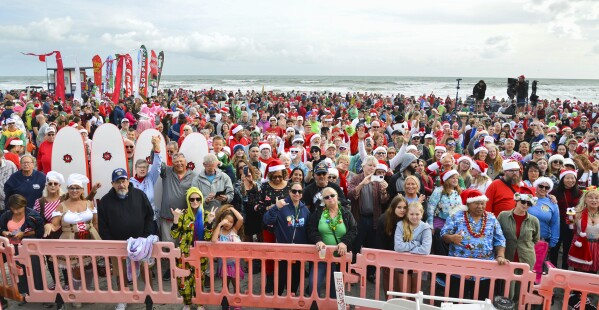 Crowds gather for the Surfing Santa event Christmas Eve morning in Cocoa Beach Fla., on Sunday, Dec. 24, 2023. The tradition started in 2009 when George Trosset and his son, George Jr., went surfing in a Santa suit and an elf costume on Christmas Eve. Friends found out about it and joined them the next year. The event grew each year and now in its 15th year the event attracts thousands and has become a fund raiser for the Florida Surf Museum and Grind for Life, an organization that provides financial assistance to cancer victims. (Malcolm Denemark/Florida Today via AP)