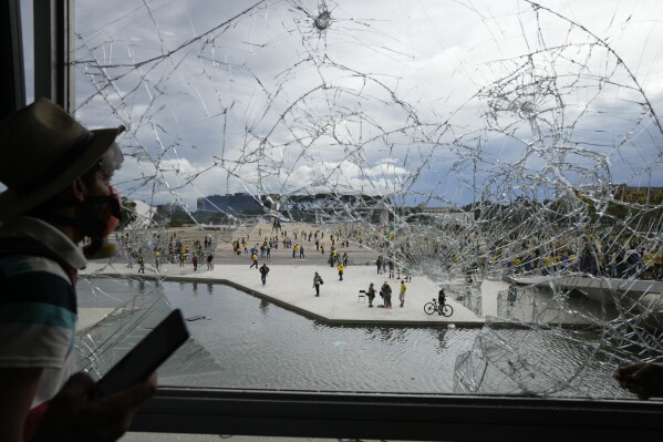 FILE - A protester, supporter of Brazil's former President Jair Bolsonaro, looks out from a shattered window of the Planalto Palace after he and many others stormed it, in Brasilia, Brazil, Jan. 8, 2023. Members of the three branches of power in Brazil say the country’s democracy and its guardrails have been restored after the trashing of the government buildings a year ago. But arrests have led supporters of the former president to say their freedom of speech is being violated and claim they are politically persecuted. (AP Photo/Eraldo Peres, File)