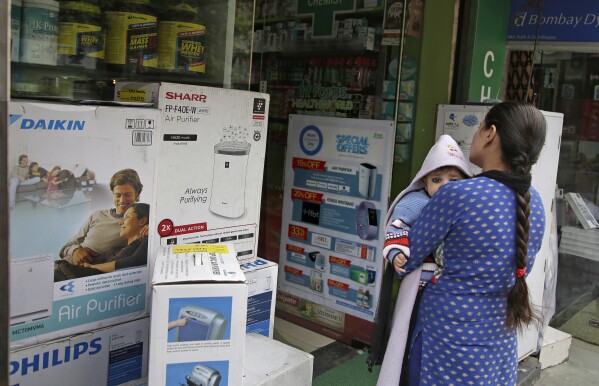 FILE- A woman carries an infant outside a shop selling air purifiers at a market place in New Delhi, India, on Nov. 14, 2017. (AP Photo/Altaf Qadri, File)
