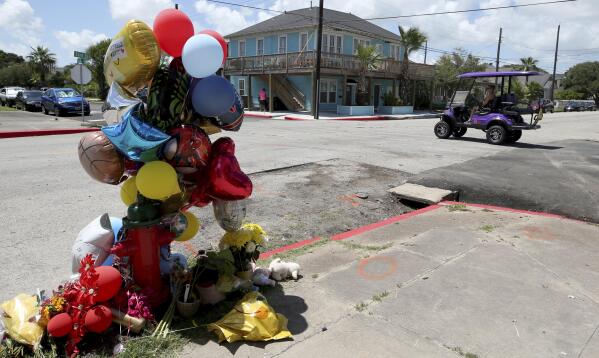 A woman driving a golf cart crosses 33rd Street at Avenue R in Galveston, Texas on Monday, Aug. 8, 2022, in Galveston, Texas, where balloons, flowers and stuffed animals have been left in memory of the victims of a fatal crash involving a golf cart and two automobiles. The four people killed in a weekend golf cart collision in Texas were a grandfather, two of his grandchildren and a niece who were visiting Galveston for a quick vacation before school began, police said. (Jennifer Reynolds/The Galveston County Daily News via AP)
