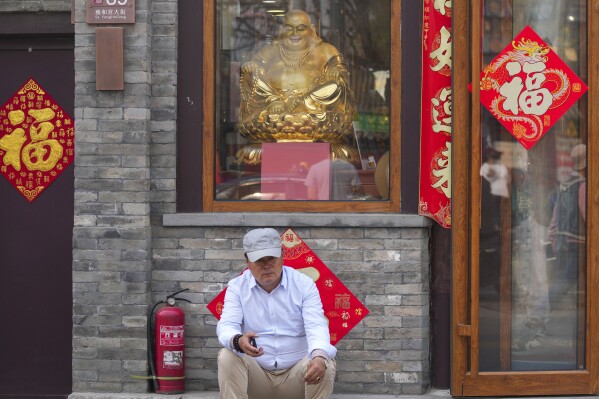 A man rests in front of a Buddha statue seen in the window of a souvenir store in Beijing, China, Friday, April 12, 2024. (AP Photo/Tatan Syuflana)
