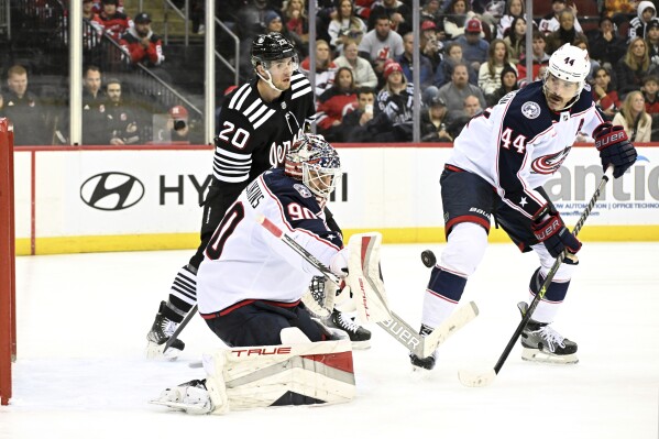 Columbus Blue Jackets goaltender Elvis Merzlikins (90) deflects the puck as defenseman Erik Gudbranson (44) and New Jersey Devils center Michael McLeod (20) look on during the second period of an NHL hockey game Friday, Nov. 24, 2023, in Newark, N.J. (AP Photo/Bill Kostroun)