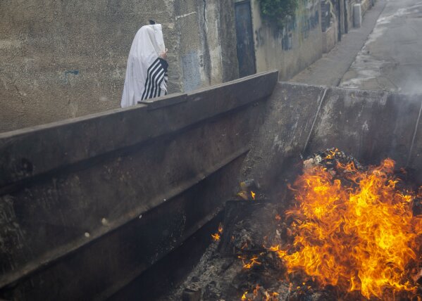 An ultra-Orthodox Jewish man wears a prayer shawl as leavened items burn in a garbage bin in final preparation for the Passover holiday in the Orthodox neighborhood of Mea Shearim in Jerusalem, Wednesday, April 8, 2020. Israeli Prime Minister Benjamin Netanyahu announced Monday a complete lockdown over the upcoming Passover holiday to control the country's coronavirus outbreak, but offered citizens some hope by saying he expects to lift widespread restrictions after the week-long festival. Passover begins on sundown Wednesday. (AP Photo/Ariel Schalit)