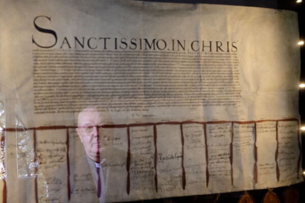 Prefect of the Archivio Apostolico Vaticano, Bishop Sergio Pagano reflects on the glass protection of the original 1530 letter kept in his office at The Vatican, Wednesday, Feb. 14, 2024, and signed and sealed by the overwhelming majority of the House of Lords that attempted to pressure Pope Clement VII into granting the divorce to King Henry VIII of England from his wife Catherine of Aragon that was famously denied. Consequently, Henry VIII made himself the head of his church, free to divorce or marry whomever he pleased. In a new book-length interview with Italian journalist Massimo Franco, “Secretum”, Pagano divulges some of the unknown or behind-the-scenes details of well-known sagas of the Holy See and its relations with the outside world over the past 12 centuries. From Napoleon’s sacking of the archive in 1810 to the Galileo affair and the peculiar conclave of 1922 that was financed almost entirely by donations from U.S. Catholics. (AP Photo/Domenico Stinellis)
