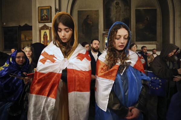 Two young women, participants in a protest against "the Russian law" and draped Georgian national flags, attend a religious service celebrating Orthodox Easter at the Trinity cathedral in Tbilisi, Georgia, late Saturday, May 4, 2024. (AP Photo/Zurab Tsertsvadze)