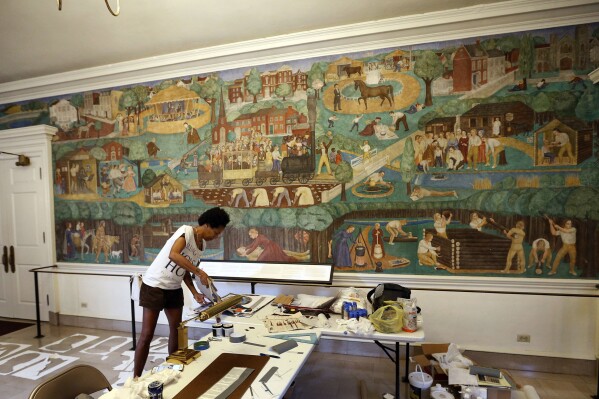 FILE - In this Aug. 15, 2018, file photo, artist Karyn Olivier creates contextual art around the University of Kentucky's controversial 1930s mural, seen on the wall in the background, in Memorial Hall in Lexington, Ky. A judge has dismissed a lawsuit that sought to stop the University of Kentucky from removing a mural that has been the object of protests for its depictions of Black people and Native Americans. But the ruling also protects the artwork. (Charles Bertram/Lexington Herald-Leader via AP, File)