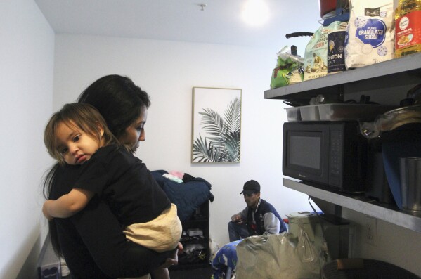 Barbara Peraza-Garcia holds her 2-year-old daughter, Frailys, by the kitchenette section of her 'micro apartment' while her partner, Franklin Peraza, browses Netflix in Seattle on Monday, March 11, 2024. (AP Photo/Manuel Valdes)