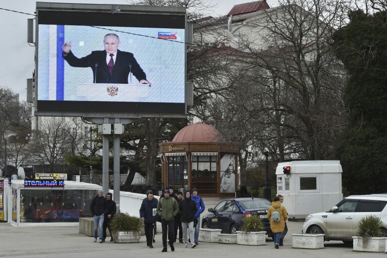 FILE - People walk in front of a tv screen showing Russian President Vladimir Putin during his annual state of the nation address in Sevastopol, Crimea, Tuesday, Feb. 21, 2023. In February 2024, Russia's central election committee said that some 112.3 million people were eligible to vote in Russia and Russian-occupied areas of Ukraine. (AP Photo, File)