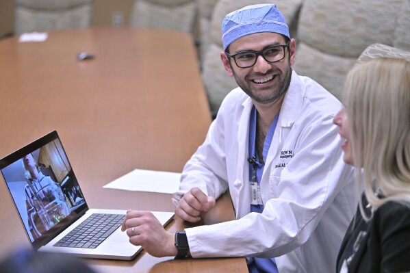 Dr. Rohaid Ali plays a video from a high school project made by his patient Alexis Bogan on Thursday, March 11, 2024, at Rhode Island Hospital in Providence, R.I. Doctors treating Bogan, whose speech was impaired by a brain tumor, used the recorded sample of her speech and a voice-cloning tool from OpenAI to recreate her previous voice. Neurosurgeon Dr. Konstantina Svokos, right, looks on. (AP Photo/Josh Reynolds)