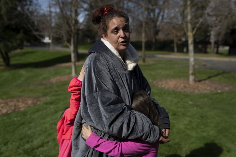 Nichole McKendrick holds her two daughters as she speaks after getting free food at Riverside Park on Thursday, March 21, 2024, in Grants Pass, Ore. McKendrick has been living out of a vehicle with her two girls. (AP Photo/Jenny Kane)