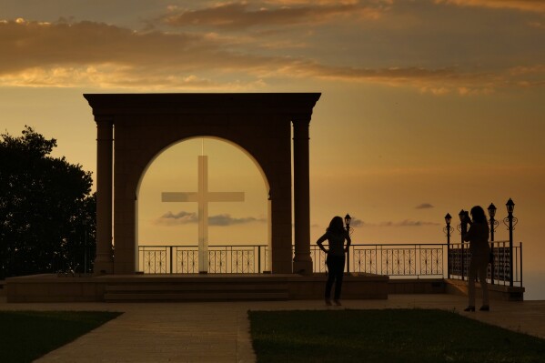 FILE - A woman uses her mobile phone to take a picture of her friend as the sun sets behind Saint Simon Church in Ghosta, in east Beirut, Lebanon, Saturday, Oct. 8, 2022. In the Middle East and North Africa, where religion is often ingrained in daily life's very fabric, rejecting faith can come with social or other repercussions, so many of the "nones," a group that includes agnostics, atheists and "nothing in particular" conceal that part of themselves, as blasphemy laws and policies are widespread in the region. (AP Photo/Hassan Ammar, File)