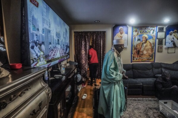Imam Omar Niass enters a living room at Bronx's Masjid Ansaru-Deen mosque, during live broadcast, left, of Ramadan prayers in Senegal at a mosque built by his grandfather, who is shown on a poster, center right, Friday March 15, 2024, at in New York. (AP Photo/Bebeto Matthews)