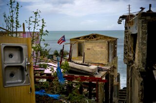 FILE - In this Oct. 5, 2017 file photo, a Puerto Rican national flag is mounted on debris of a damaged home in the aftermath of Hurricane Maria in the seaside slum La Perla, San Juan, Puerto Rico. An independent investigation ordered by Puerto Rico’s government estimates that nearly 3,000 people died as a result of Hurricane Maria. The findings issued Tuesday, Aug. 28, 2018, by the Milken Institute School of Public Health at George Washington University contrast sharply with the official death toll of 64. (AP Photo/Ramon Espinosa)