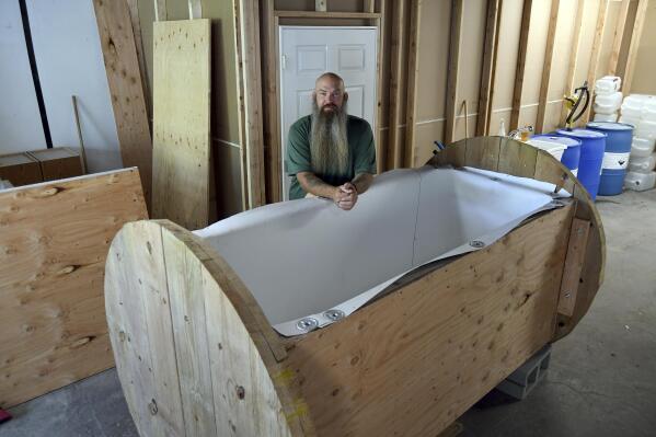 In this Wednesday, Aug. 11, 2021, photo, Seth Viddal, who co-owns The Natural Funeral, stands behind a nearly completed human body composting vessel in Arvada, Colo. On Sept. 7, Colorado became the second state after Washington to allow human body composting, and Oregon will allow the practice beginning next July. The vessel will be packed with wood chips and straw and will be able to compost a body in six months. About the size of a standard grave, the rectangular insulated wooden box is lined with waterproof roofing material and packed with wood chips and straw. Two large spool wheels on either end allow it to be rolled across the floor, providing the oxygenation, agitation and absorption required for a body to compost. (AP Photo/Thomas Peipert)