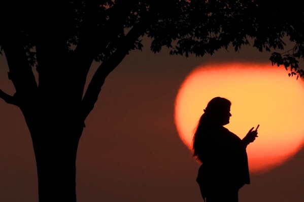 A woman looks at her phone while watching the sun set in Kansas City, Mo., on Sunday, Aug. 20, 2023. (AP Photo/Charlie Riedel, File)