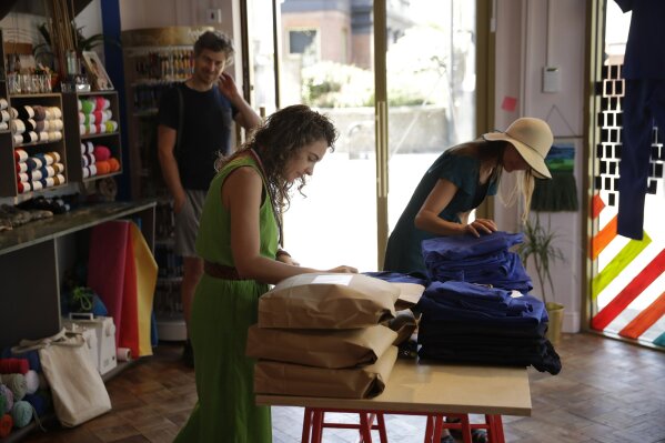 Textile artist Brooke Dennis, centre, who is originally from New Zealand, receives scrubs made for NHS (National Health Service) staff to wear during the coronavirus outbreak from a volunteer, right, at Brooke's textiles and craft studio called Make Town, in east London, Thursday, April 23, 2020, as part of the Scrub Hub network of voluntary community groups.  The Scrub Hub just wanted to help, but they created a movement with some 70 hubs employing the skills of more than 2,200 volunteers all over the nation responding to the coronavirus pandemic, with a template for PPE, a pattern that so far has made more than 3,800 sets of scrubs for healthcare workers. (AP Photo/Matt Dunham)