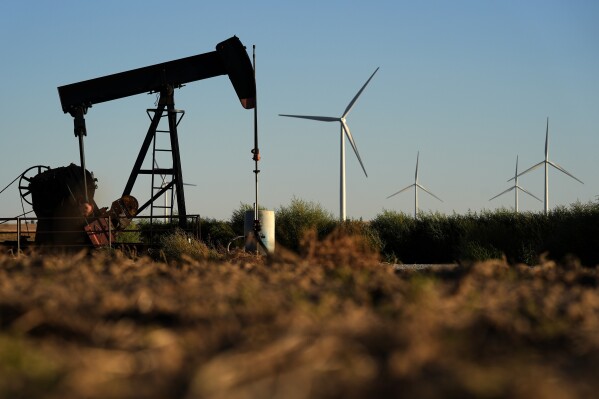 Pumpjacks operate in the foreground while wind turbines at the Buckeye Wind Energy wind farm rise in the distance, Sept. 30, 2024, near Hays, Kan. (AP Photo/Charlie Riedel, File)