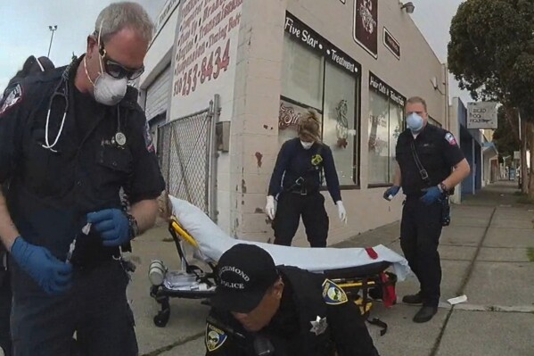 In this image from Richmond Police Department body-camera video, a paramedic, left, holds a syringe as he prepares to inject Ivan Gutzalenko, 47, with the sedative midazolam in Richmond, Calif., on March 10, 2021. When the paramedic returned three minutes after the injection, Gutzalenko lay motionless. He was declared dead at a hospital. An investigation by The Associated Press found that at least 16 people died in California in 2012-2021 following physical encounters with police during which medical personnel also injected them with a powerful sedative. (Richmond Police Department via AP)