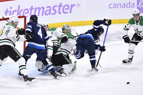 Dallas Stars goaltender Jake Oettinger, center, upends Winnipeg Jets' Vladislav Namestnikov, second from right, as Stars' Mason Marchment (27) looks for the loose puck during the first period of an NHL hockey match in Winnipeg, Manitoba, on Tuesday, Nov. 28, 2023. (Fred Greenslade/The Canadian Press via AP)