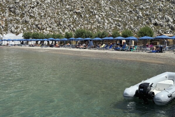 People sit on the beach of Agios Nikolaos from where British doctor and television presenter Michael Mosley, is believed to have set out, on the southeastern Aegean Sea island of Symi, Greece, Friday, June 7, 2024. Greek police say an ongoing major search and rescue operation on the small eastern Aegean island of Symi has still not located British doctor and television presenter Michael Mosley, who went missing on Wednesday afternoon after reportedly going for a walk. (AP Photo/Antonis Mystiloglou)