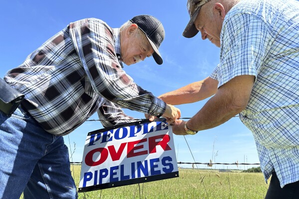 Gaylen Dewing, left, and Marvin Abraham affix a sign to a roadside fence east of Bismarck, N.D., on Tuesday, Aug. 15, 2023. The sign reads "People over pipelines," and is in opposition to Summit Carbon Solutions' proposed five-state, 2,000-mile pipeline network to carry carbon dioxide emissions from dozens of Midwestern ethanol plants to North Dakota for permanent storage deep underground. (AP Photo/Jack Dura)