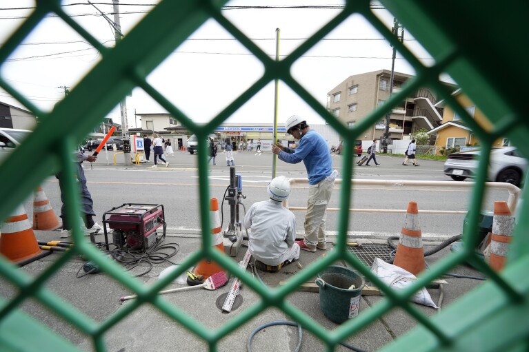 Workers set up a barricade near the Lawson convenience store, background, Tuesday, April 30, 2024, at Fujikawaguchiko town, Yamanashi Prefecture, central Japan. The town of Fujikawaguchiko, known for a number of popular photo spots for Japan's trademark of Mt. Fuji, on Tuesday began to set up a huge black screen on a stretch of sidewalk to block view of the mountain in a neighborhood hit by a latest case of overtourism in Japan. (AP Photo/Eugene Hoshiko)