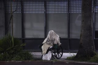A homeless person sits on a wheelchair under rainy weather on Sunset Blvd., in the Echo Park neighborhood of Los Angeles Monday, April 6, 2020. One population is particularly vulnerable to contracting and spreading the coronavirus: the homeless. Officials have vowed repeatedly to get them indoors, but testing shortages and bureaucratic wrangling are making it difficult. Relatively few of California's 150,000 homeless population have been moved into individual quarters. It's unclear how many even have the highly contagious virus. It's a problem playing out nationwide and it's unclear how many may even have coronavirus. (AP Photo/Damian Dovarganes)