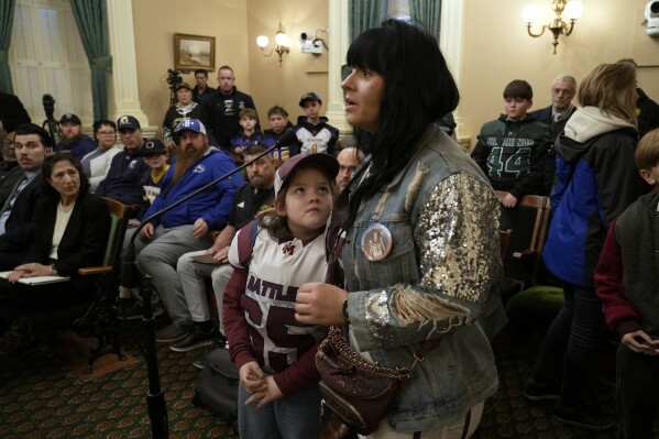 Bruce Bertram, 7, who plays tackle football for the Union Mine Junior Rattlers looks up at his mother, Ashley Bertram, as she speaks against a proposed California bill that would ban children under 12 from playing tackle football, during a legislative committee hearing in Sacramento, Calif., Wednesday, Jan. 10, 2024. (AP Photo/Rich Pedroncelli)