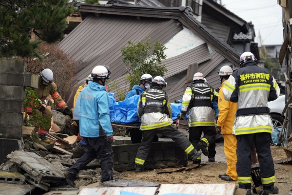 Feuerwehrleute und Rettungskräfte tragen eine Leiche mit einem blauen Laken, die aus einem eingestürzten Haus geborgen wurde, das nach einem starken Erdbeben in Suzu, Präfektur Ishikawa, am Mittwoch, 3. Januar 2024, eingestürzt war. (AP Photo/Hiro Komai)