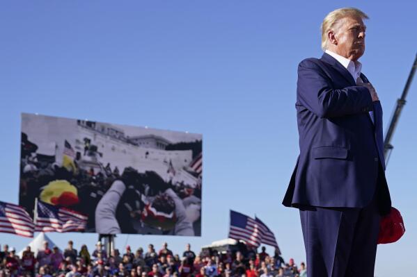 FILE - As footage from the Jan. 6, 2021, insurrection at the U.S. Capitol is displayed in the background, former President Donald Trump stands while a song, "Justice for All," is played during a campaign rally at Waco Regional Airport, Saturday, March 25, 2023, in Waco, Texas. The tune, “Justice for All,” is the Star-Spangled Banner and it was sung by a group of defendants jailed over their alleged roles in the January 2021 insurrection. The national anthem is overlaid with Trump reciting the Pledge of Allegiance. (AP Photo/Evan Vucci, File)