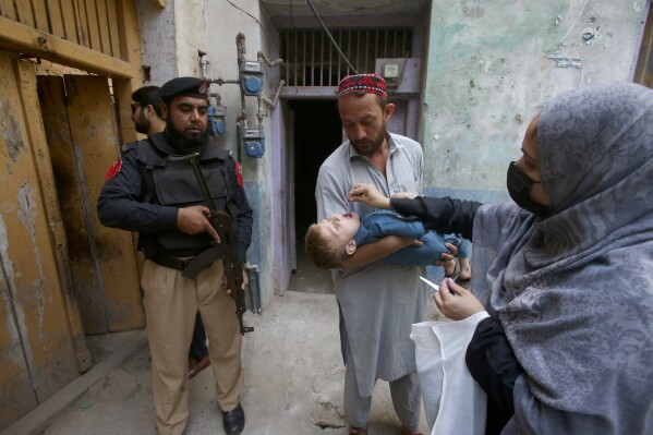 A police officer stands guard as a health worker administers a polio vaccine to a child at a neighborhood of Peshawar, Pakistan, Monday, Oct. 2, 2023. (AP Photo/Muhammad Sajjad)