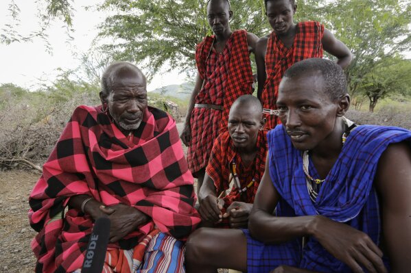 In this photo taken on Thursday, April 9, 2020, Maasai Village elder Letaari Salaash, left, sits with younger Maasai warriors at their village in Kajiado County in Kenya. The Maasai, a semi-nomadic indigenous group in Kenya and Tanzania, have been forced to halt important rituals that bring clans together due to the coronavirus, including the graduation of warriors into young men who can marry and own property. (AP Photo/Khalil Senosi)