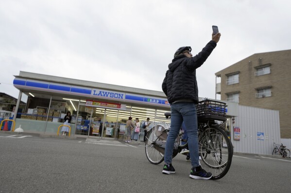 Ein Tourist macht am Dienstagabend, dem 30. April 2024, ein Selfie im Lawson Department Store, einem beliebten Fotospot mit einem malerischen Blick auf den Berg Fuji im Hintergrund, in der Stadt Fujikawaguchiko, Präfektur Yamanashi, Zentraljapan.  Die Stadt Fujikawaguchiko, die für eine Reihe beliebter Drehorte für die japanische Marke „Mount Fuji“ bekannt ist, hat am Dienstag damit begonnen, eine riesige schwarze Leinwand auf dem Bürgersteig zu platzieren, um den Blick auf den Berg in einem Viertel zu versperren, das von Japans jüngstem Fall von Übertourismus heimgesucht wurde.  (AP Photo/Eugene Hoshiko)