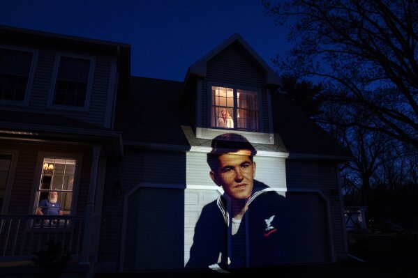 An image of veteran Stephen Kulig is projected onto the home of his daughter, Elizabeth DeForest, as she looks out the window of a spare bedroom as her husband, Kevin, sits downstairs in Chicopee, Mass., Sunday, May 3, 2020. Kulig, a U.S. Navy veteran and resident of the Soldier's Home in Holyoke, Mass., died from the COVID-19 virus at the age of 92. After saying goodbye to her father for the last time in person, Elizabeth slept in the spare bedroom upstairs for two weeks as a precaution against possibly infecting her husband. Seeking to capture moments of private mourning at a time of global isolation, the photographer used a projector to cast large images of veterans on to the homes as their loved ones are struggling to honor them during a lockdown that has sidelined many funeral traditions. (AP Photo/David Goldman)