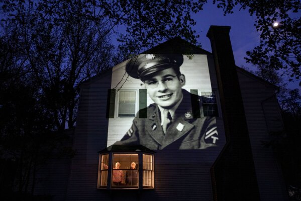 An image of veteran James Sullivan is projected onto the home of his son, Tom Sullivan, left, as he looks out a window with his brother, Joseph Sullivan, in South Hadley, Mass., Monday, May 4, 2020. Sullivan, a U.S. Army WWII veteran and resident of the Soldier's Home in Holyoke, Mass., died from the COVID-19 virus four days shy of his 100th birthday. Seeking to capture moments of private mourning at a time of global isolation, the photographer used a projector to cast large images of veterans on to the homes as their loved ones are struggling to honor them during a lockdown that has sidelined many funeral traditions. (AP Photo/David Goldman)