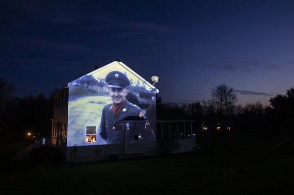 An image of veteran Chester LaPlante is projected onto the home of his son, Randy LaPlante, as he looks out a window with his wife, Nicole, and their sons, Evan and Blake, at their home in Amsterdam, N.Y., Tuesday, May 5, 2020. LaPlante, a U.S. Army veteran and resident of the Soldier's Home in Holyoke, Mass., died from the COVID-19 virus at the age of 78. Seeking to capture moments of private mourning at a time of global isolation, the photographer used a projector to cast large images of veterans on to the homes as their loved ones are struggling to honor them during a lockdown that has sidelined many funeral traditions. (AP Photo/David Goldman)