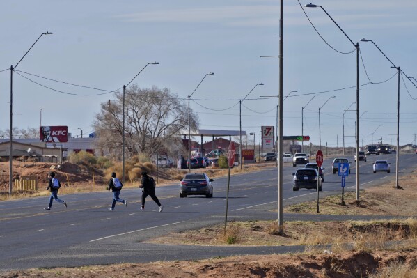 Teens cross Main Street on the Navajo Nation, Monday, March 4, 2024, in Tuba City, Ariz., just north of U.S. 160. The highway is the de facto border between the Navajo and Hopi Indian reservations and two time zones. Mind-bending time calculations is what people in the largest Native American reservation in the U.S. have to endure every March through November. (AP Photo/Matt York)