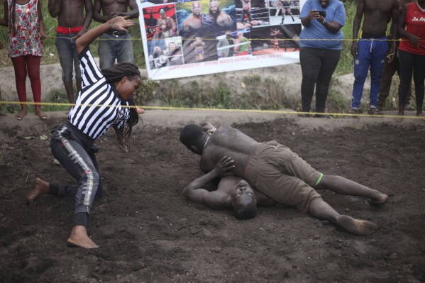 CORRECTS THE YEAR TO 2024 - Ugandan youths perform an amateur wrestling tangle in the soft mud in Kampala, Uganda Wednesday, March 20, 2024. The open-air training sessions, complete with an announcer and a referee, imitate the pro wrestling contests the youth regularly see on television. While a pair tangles inside the ring, made with bamboo poles strung with sisal rope, others standing ringside cheer feints and muscular shows of strength. (AP Photo/Patrick Onen)